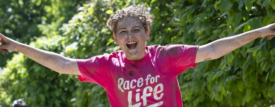 A young boy is having fun taking part in a Pretty Muddy Kids race. He is wearing a pink T-shirt and covered in muddy water.