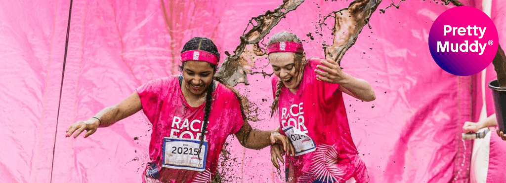 Two women smiling wearing pink race for life tshirts getting mud thrown over them at pretty muddy event.